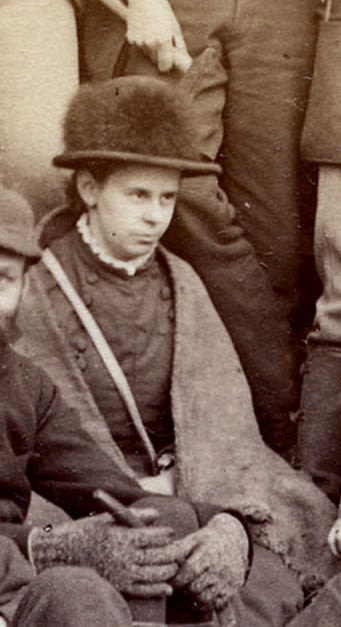 Photograph of women seated holding a geological hammer. She is wearing a hat and has a bag over her shoulder.
