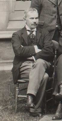Photograph of a man sitting down outside. He has his arms folded. He is wearing a suit jacket and tie.