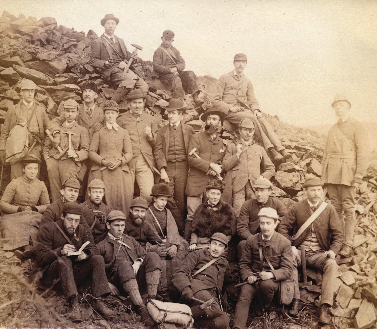 Sepia photograph of a group of students on a field trip in Wales. The students are wearing hats and holding hammers.