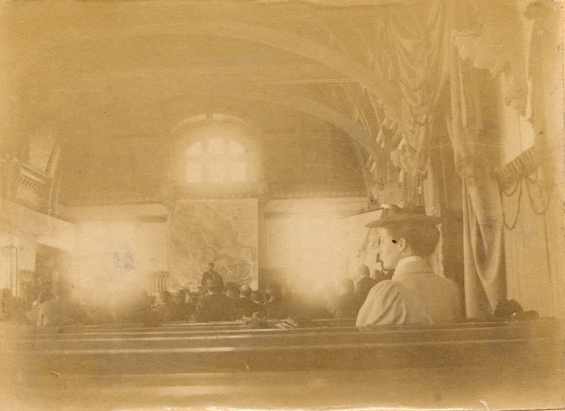 Sepia photograph of a lecture. A map can be seen behind the lecturer. A woman is sitting at the back of the room.