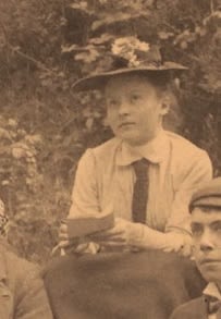 Photograph of woman outside holding a notebook and pencil. She is wearing a hat with flowers on it.
