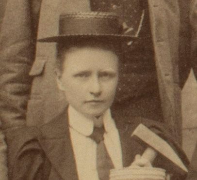 Photograph of a woman holding a geological hammer. She is wearing a hat and tie.