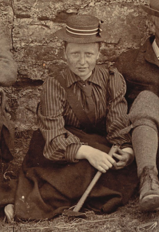 Photograph of a seated woman wearing a hat. She is holding a geologicla hammer.