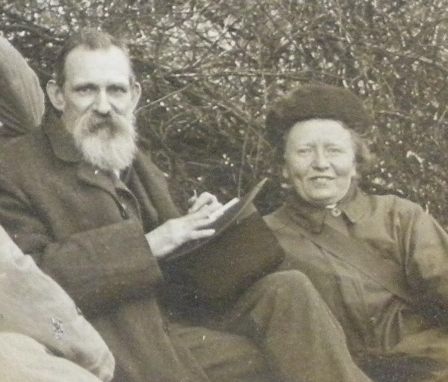 Two adults sitting down outside. The man is holding a hat. The lady is wearing a hat. Both are facing the camera and smiling.