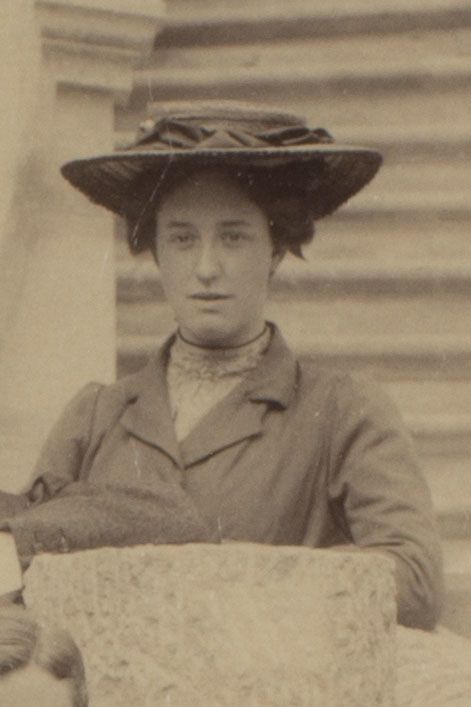 Photograph of woman standing by some steps. She is wearing a large hat, and is facing the camera.