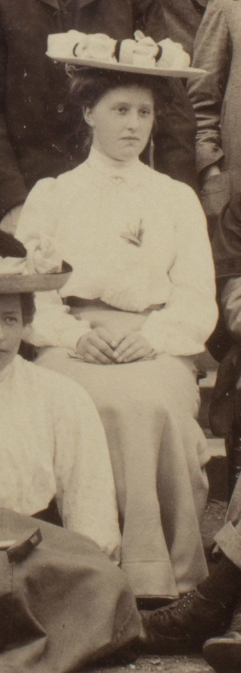 Formal photograph of woman seated outside. She is wearing a hat and has her hands in her lap.