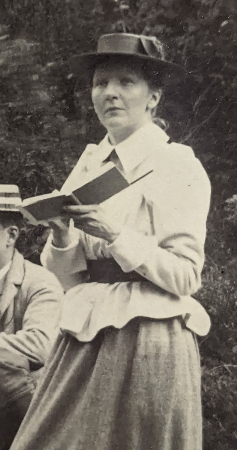 Photograph of a woman standing outside, wearing a hat. She is holding a notebook and pencil.