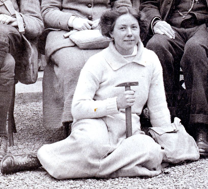 Photograph of woman seated outside holding a geological hammer. She has a small bag to her left hand side.