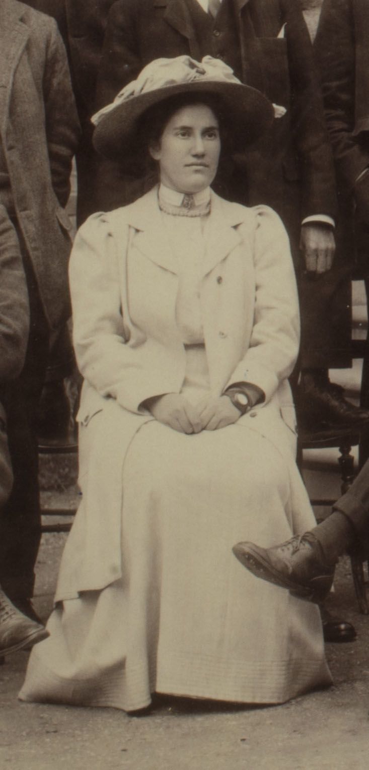 Formal photograph of woman seated outside. She is wearing a hat and has her hands clasped.