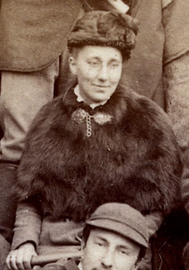 Photograph of a woman seated outside holding a geological hammer. She is wearing a fur shawl and hat.
