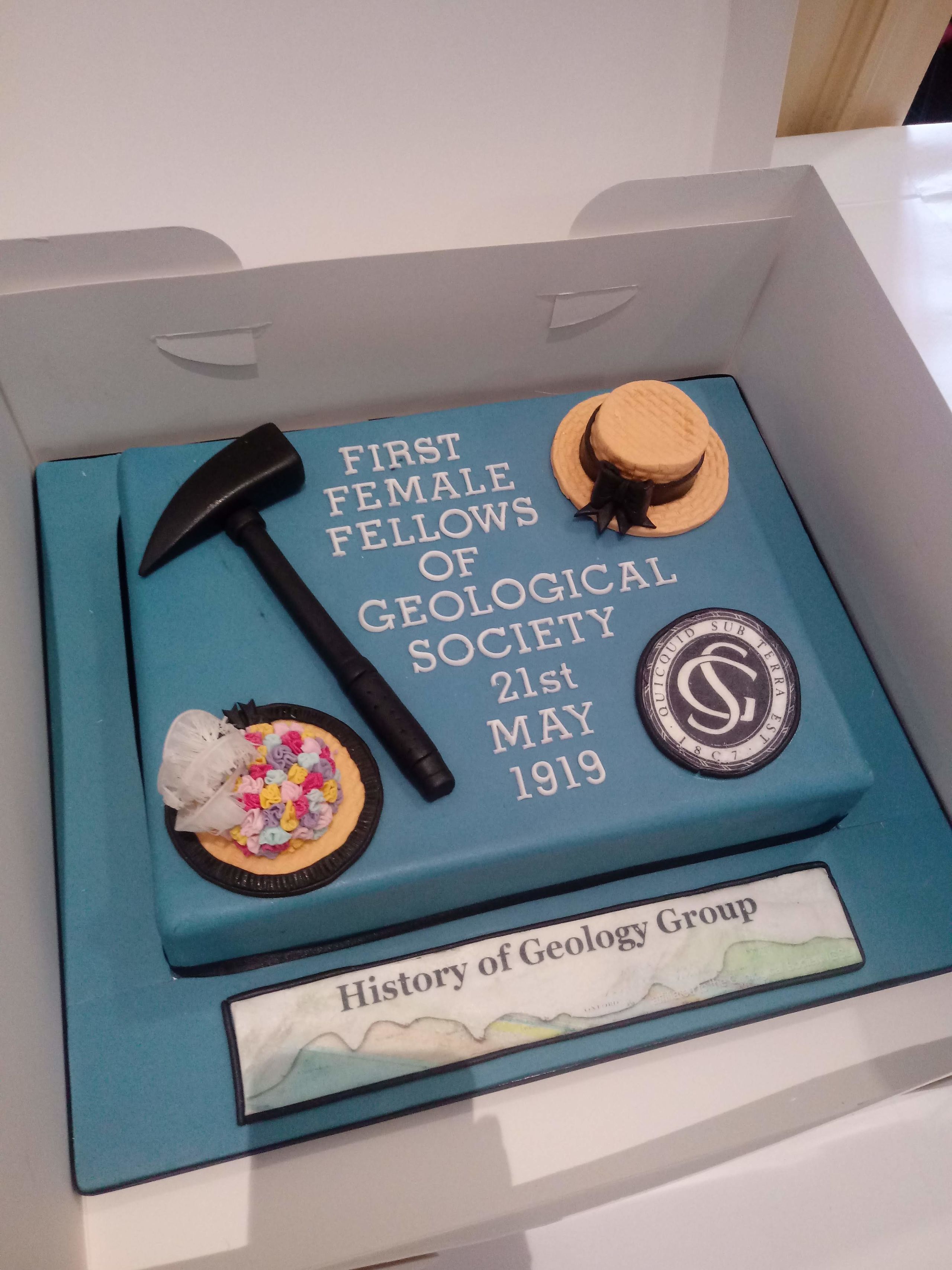 Photograph of a cake. It is square with sugar paste hats and a hammer, and text. 