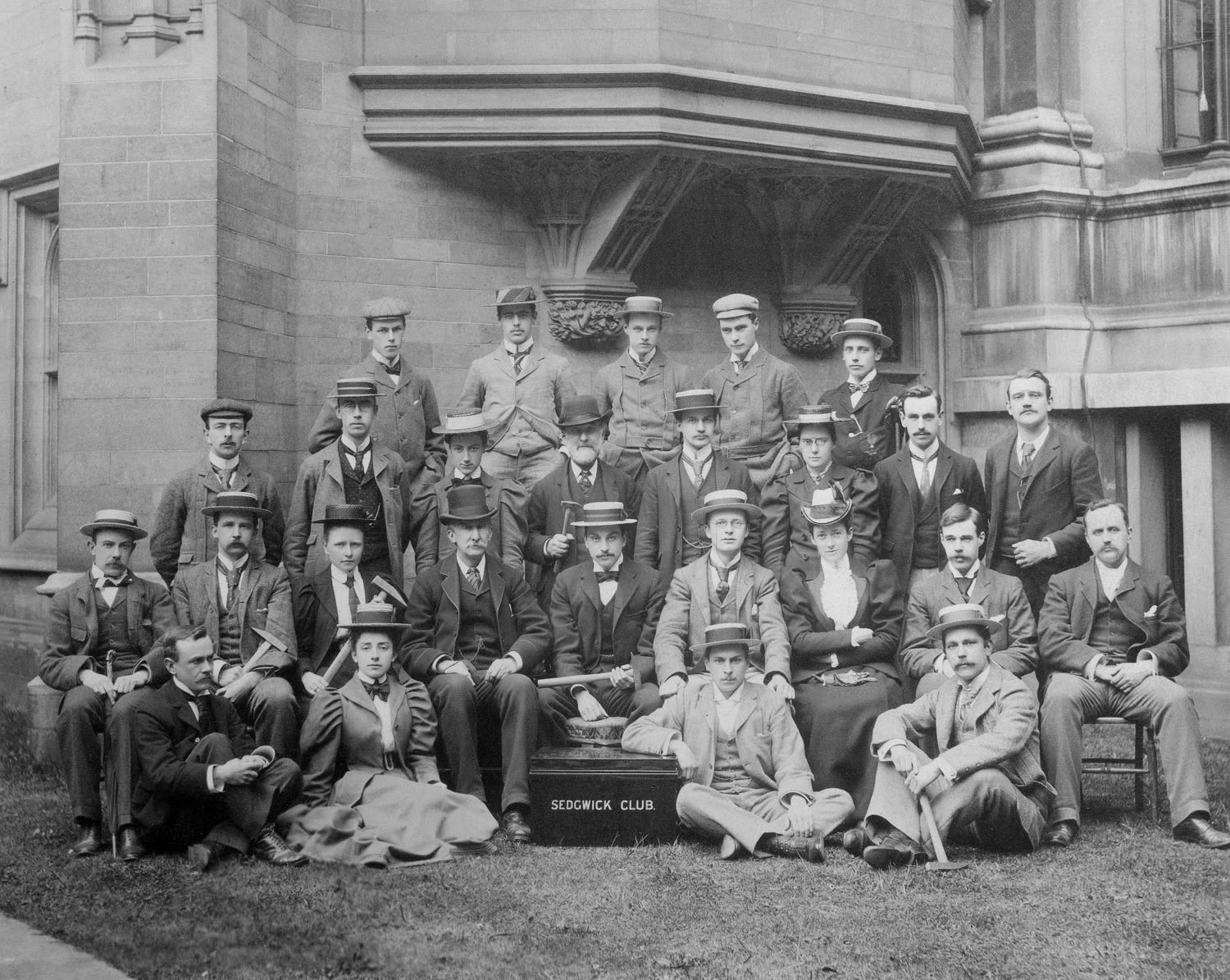 Black and white formal photograph outside. Students are wearing hats and holding hammers. There is a large black tin in the foreground. 