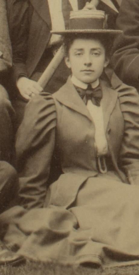 Formal photograph of woman seated outside. She is wearing a hat and a bow tie. A large geological hammer is being held behind her.