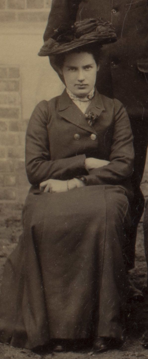 Formal photograph of woman seated outside. She is wearing a hat and has her arms crossed.